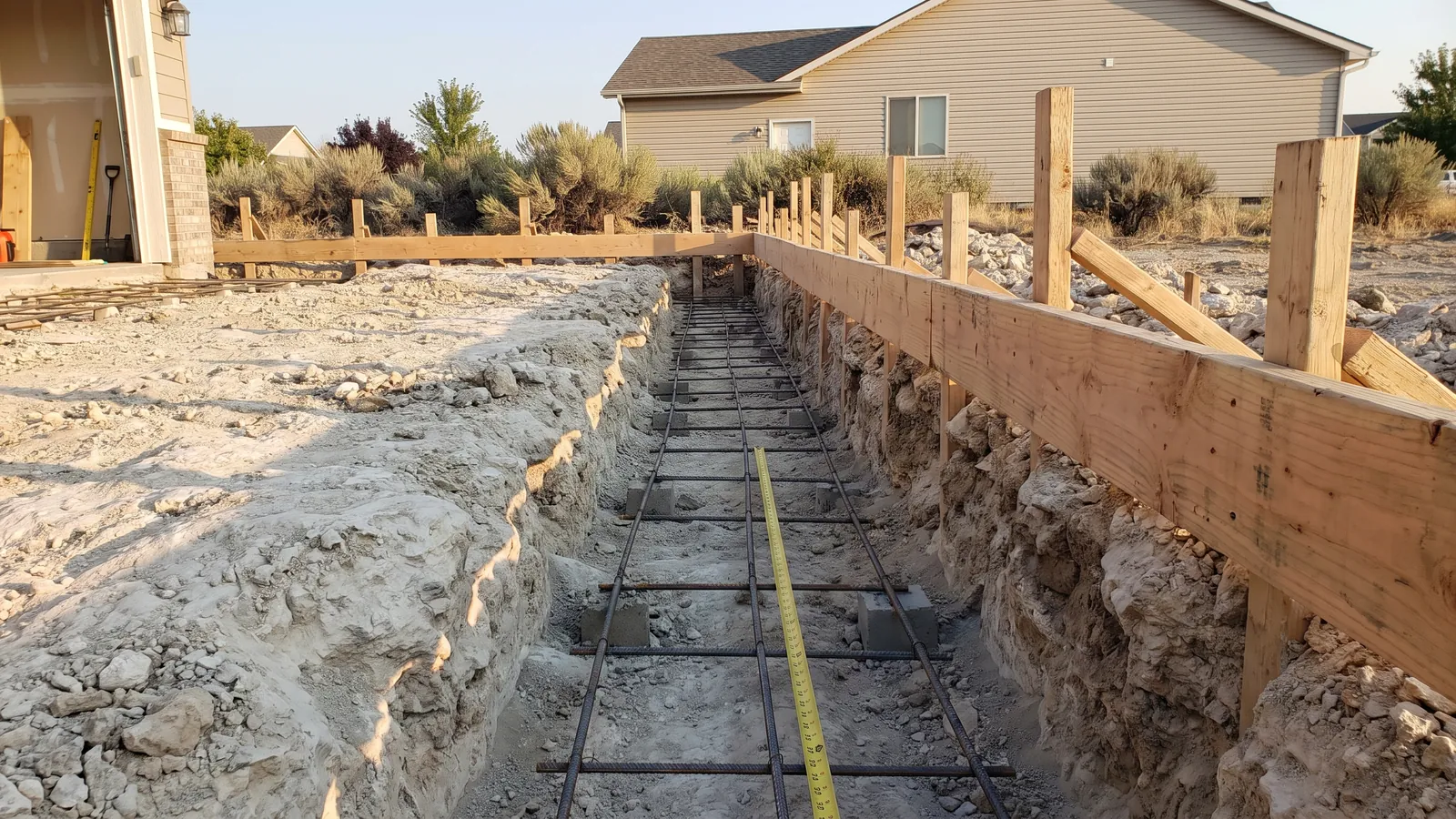 Footing trench cut through pale cemented hardpan to frost depth, with rebar on chairs and formwork for a garage addition in Idaho